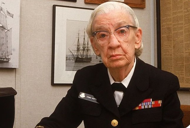 Grace Hopper in her US Navy uniform, seated in an office in front of framed pictures of sailing ships