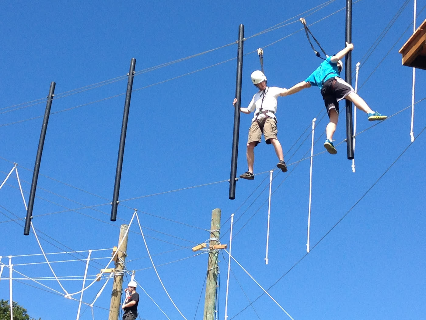 Two people on a high ropes course, one reaching out and holding the other’s arm while both stand on cables high above the ground